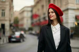 mode femme portrait sourire avec les dents permanent sur le rue dans le ville Contexte dans élégant vêtements rouge lèvres et rouge béret, voyage, cinématique couleur, rétro ancien style, Urbain mode mode de vie. photo