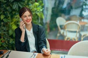 une femme blogueur touristique est assis dans une café à une table avec une téléphone dans sa mains parlant sur le téléphone, mobile les communications et le l'Internet sur une printemps voyage, affaires appel, free-lance travail en ligne, sourire photo