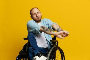 une homme dans une fauteuil roulant regards à le caméra, avec tatouages sur le sien bras est assis sur une Jaune studio arrière-plan, le concept de santé est une la personne avec handicapées photo