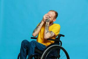 une homme dans une fauteuil roulant sourire regards à le caméra dans une T-shirt avec tatouages sur le sien bras est assis sur une bleu studio arrière-plan, une plein vie, une réel la personne photo