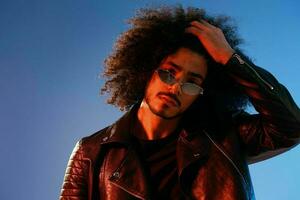 portrait de une élégant homme avec frisé cheveux sur une bleu Contexte multinational, coloré lumière, branché lunettes, moderne concept. photo