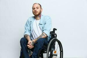 une homme dans une fauteuil roulant regards pensivement à le caméra, copie espace, avec tatouages sur le sien bras est assis sur une gris studio arrière-plan, le concept de santé une la personne avec handicapées, une réel la personne photo