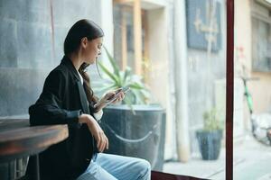 élégant femelle blogueur est assis dans une café avec une téléphone dans sa mains, mobile les communications et l'Internet à l'étranger, vidéo appel, en ligne free-lance travail photo