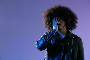 portrait de mode homme avec frisé cheveux sur bleu Contexte multinational, coloré lumière, noir cuir veste s'orienter, moderne concept. photo