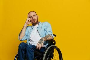 une homme dans une fauteuil roulant problèmes avec le musculo-squelettique système regards à le caméra pensivement avec tatouages sur le sien bras est assis sur une Jaune studio arrière-plan, santé concept une la personne avec handicapées photo