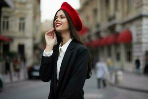 mode femme portrait sourire avec les dents permanent sur le rue dans le ville Contexte dans élégant vêtements rouge lèvres et rouge béret, voyage, cinématique couleur, rétro ancien style, Urbain mode mode de vie. photo