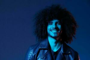 portrait de mode homme avec frisé cheveux sur bleu Contexte multinational, coloré lumière, noir cuir veste s'orienter, moderne concept. photo