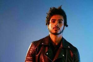 portrait de une élégant homme avec frisé cheveux sur une bleu Contexte multinational, coloré lumière, noir cuir veste s'orienter, moderne concept. photo