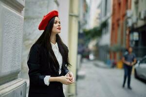 une femme permanent près une mur dans le ville portant une élégant veste et une rouge béret avec rouge lèvres, en voyageant. photo