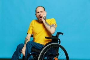 une homme dans une fauteuil roulant surpris et ouvert bouche dans une T-shirt avec tatouages sur le sien bras est assis sur une bleu studio arrière-plan, une plein vie, une réel la personne photo