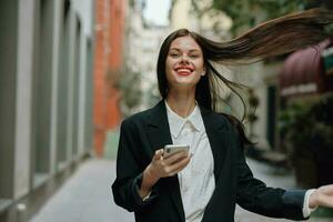 affaires brunette femme avec rouge lèvres sourire avec les dents avec une téléphone dans sa mains, blanc chemise et noir veste mode sur le rue, été voyage, vacances dans le ville touristique pigiste photo