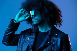 portrait de mode homme avec frisé cheveux avec élégant des lunettes sur bleu Contexte multinational, coloré lumière, noir cuir veste s'orienter, moderne concept. photo