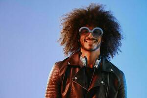 portrait de une élégant homme avec frisé cheveux avec des lunettes et écouteurs sur une bleu Contexte multinational, coloré lumière, noir cuir veste s'orienter, moderne concept. photo