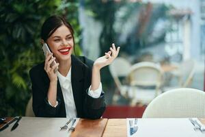 femme blogueur touristique est assis dans une café à une table avec une téléphone dans sa mains parlant sur le téléphone, mobile les communications et le l'Internet sur une printemps voyage, vidéo appel, free-lance travail en ligne, sourire photo