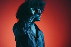 portrait de mode homme avec frisé cheveux sur rouge Contexte avec élégant lunettes, multiculturel, coloré lumière, noir cuir veste s'orienter, moderne concept. photo