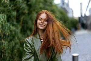 mode femme dans le ville à l'extérieur posant avec une sourire contre bâtiments et des arbres avec rouge longue en volant cheveux dans le venteux temps photo