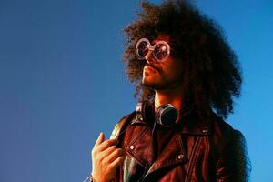 portrait de une élégant homme avec frisé cheveux avec des lunettes et écouteurs sur une bleu Contexte multinational, coloré lumière, noir cuir veste s'orienter, moderne concept. photo