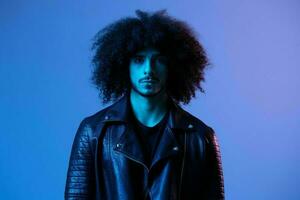 portrait de mode homme avec frisé cheveux sur bleu Contexte multinational, coloré lumière, noir cuir veste s'orienter, moderne concept. photo