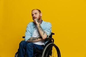 une homme dans une fauteuil roulant problèmes avec le musculo-squelettique système regards à le caméra pensivement avec tatouages sur le sien bras est assis sur une Jaune studio arrière-plan, santé concept une la personne avec handicapées photo