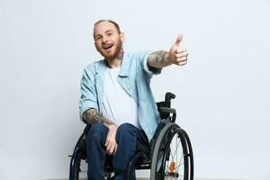 une homme dans une fauteuil roulant regards à le caméra spectacles une les pouces en haut, bonheur, avec tatouages sur le sien mains est assis sur une gris studio arrière-plan, santé concept homme avec handicapées photo