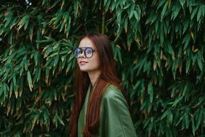 magnifique femme dans élégant vêtements portant des lunettes avec rouge cheveux sourire posant dans de face de une bambou arbre photo