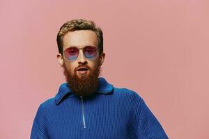 portrait de une rousse homme portant des lunettes de soleil souriant et dansant, écoute à la musique sur une rose Contexte. branché avec une barbe, bonheur doigt montrer du doigt. photo