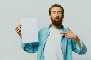 portrait de un adulte Masculin sur une gris Contexte avec une blanc feuille de papier dans le sien mains pour votre conception et texte, mise en page, copie espace, espace pour texte, doigt montrer du doigt photo