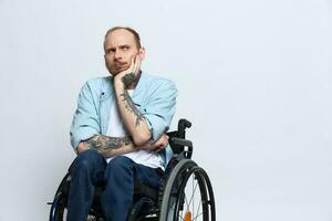 une homme dans une fauteuil roulant regards à le caméra arrogance, avec tatouages sur le sien bras est assis sur une gris studio arrière-plan, le concept de santé est une la personne avec handicapées photo