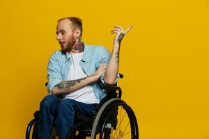 une homme dans une fauteuil roulant problèmes avec le musculo-squelettique système regards à le caméra spectacles une doigt sur, avec tatouages sur le sien mains est assis sur une Jaune studio arrière-plan, santé concept homme avec handicapées photo