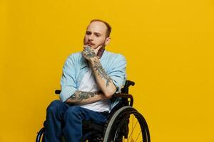 une homme dans une fauteuil roulant problèmes avec le musculo-squelettique système regards à le caméra pensivement avec tatouages sur le sien bras est assis sur une Jaune studio arrière-plan, santé concept une la personne avec handicapées photo