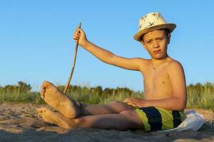 garçon dans une Soleil chapeau et nager short est relaxant séance sur le plage photo