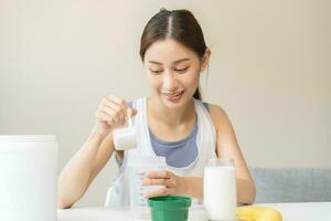 régime repas remplacement pour poids perte, sourire asiatique Jeune femme, fille dans tenue de sport, main dans en portant scoop fabrication protéine secouer, boisson supplément pour muscle après faire des exercices à maison. en bonne santé corps se soucier. photo