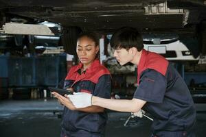 deux professionnel automobile ouvrier les partenaires discuter mécanique vérifier châssis de levé électrique voiture, réparer et réparation à entretien garage, et expert un service affaires travaux industrie profession. photo