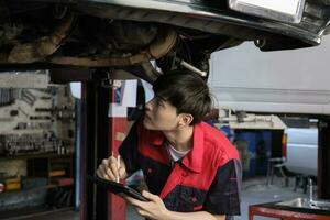 professionnel Jeune asiatique Masculin moteur mécanicien inspecte châssis de électrique voiture levé par chariot élévateur jack pour réparation à garage, automobile entretien un service travaux industrie Occupation entreprise. photo