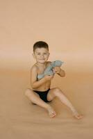 une enfant garçon dans caleçon détient une doux ours jouet dans le sien mains, séance sur une beige Contexte dans le studio et souriant photo