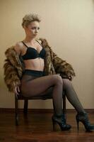 attrayant blond modèle avec collants, noir soutien-gorge et fourrure manteau posant provocateur sur chaise. mode portrait de sensuel blond, studio tir. sensuel femelle dans noir lingerie posant contre mur. photo