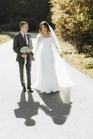 un inhabituel, pittoresque marcher de jeunes mariés sur leur mariage journée. une Jeune homme dans une costume et une magnifique mince la mariée dans une mariage robe sur le route. le la mariée est souriant tandis que en portant sa voile. photo