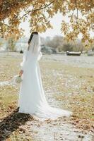 impressionnant la mariée dans une longue mariage robe avec une magnifique bouquet dans main et avec charmant sourire. en dessous de le arbre dans le magnifique le coucher du soleil lumière dans le parc. photo