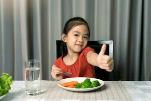 mignonne peu fille montrant pouce montrant en mangeant en bonne santé des légumes. photo