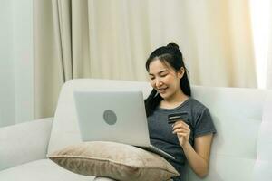 femme asiatique tenant une carte de crédit pour faire des achats en ligne tout en étant assise sur le canapé pendant les vacances à la maison. photo