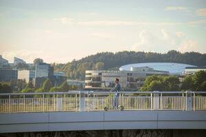 Vilnius, Lituanie 03 06 2022 Jeune homme sur scooter conduite le long de une blanc pont plus de rivière avec vert des arbres et Bureau bâtiments sur Contexte photo