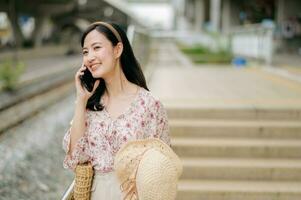 asiatique Jeune femme voyageur avec tissage chapeau en utilisant une mobile téléphone à côté de chemin de fer train station dans Bangkok. périple voyage mode de vie, monde Voyage explorateur ou Asie été tourisme concept. photo