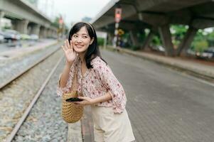 asiatique Jeune femme voyageur avec tissage panier en utilisant une mobile téléphone à côté de chemin de fer train station dans Bangkok. périple voyage mode de vie, monde Voyage explorateur ou Asie été tourisme concept. photo