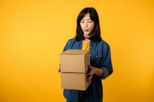 portrait asiatique Jeune femme portant Jaune T-shirt et denim chemise en portant parcelle boîte isolé sur Jaune studio arrière-plan, livraison courrier et livraison un service concept. photo