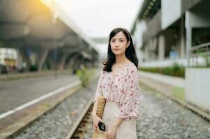 Jeune asiatique femme voyageur avec tissage panier content souriant à la recherche à une caméra à côté de train chemin de fer. périple voyage mode de vie, monde Voyage explorateur ou Asie été tourisme concept. photo