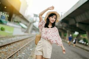 Jeune asiatique femme voyageur avec tissage panier content souriant à la recherche à une caméra à côté de train chemin de fer. périple voyage mode de vie, monde Voyage explorateur ou Asie été tourisme concept. photo