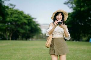 portrait de asiatique Jeune femme voyageur avec tissage chapeau et panier et une caméra sur vert Publique parc la nature Contexte. périple voyage mode de vie, monde Voyage explorateur ou Asie été tourisme concept. photo