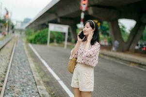 asiatique Jeune femme voyageur avec tissage panier en utilisant une mobile téléphone à côté de chemin de fer train station dans Bangkok. périple voyage mode de vie, monde Voyage explorateur ou Asie été tourisme concept. photo