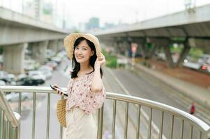 Jeune asiatique femme voyageur avec tissage panier en utilisant mobile téléphone et permanent sur dépasser avec chemin de fer Contexte. périple voyage mode de vie, monde Voyage explorateur ou Asie été tourisme concept. photo