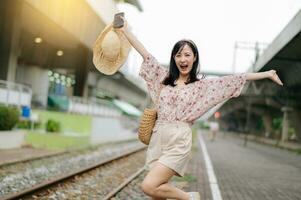 Jeune asiatique femme voyageur avec tissage panier content souriant à la recherche à une caméra à côté de train chemin de fer. périple voyage mode de vie, monde Voyage explorateur ou Asie été tourisme concept. photo